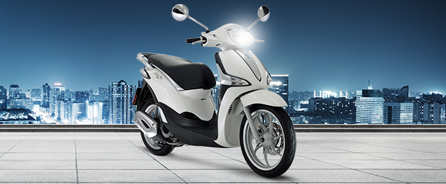 Weiße Piaggio Medley Roller auf einem Platz am Abend vor einer Stadtkulisse.